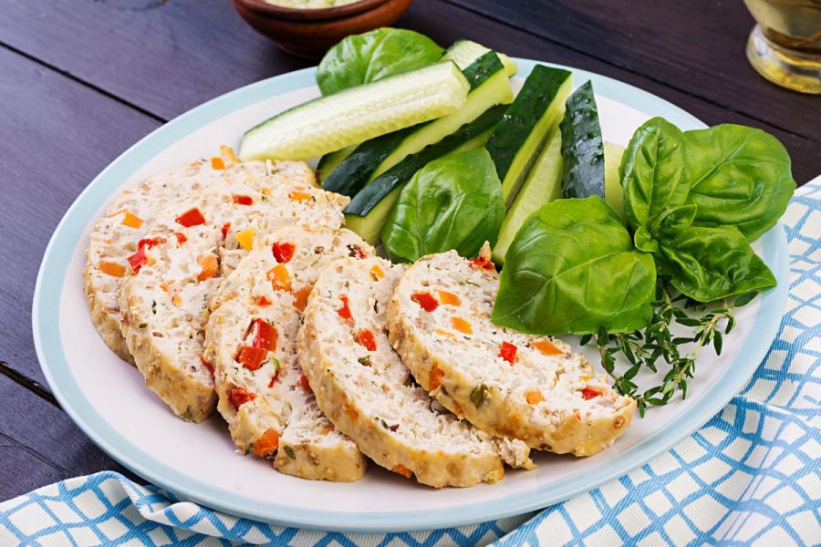 Slices of chicken meatloaf with vegetables in a plate with cucumber sticks and baby spinach on the side.